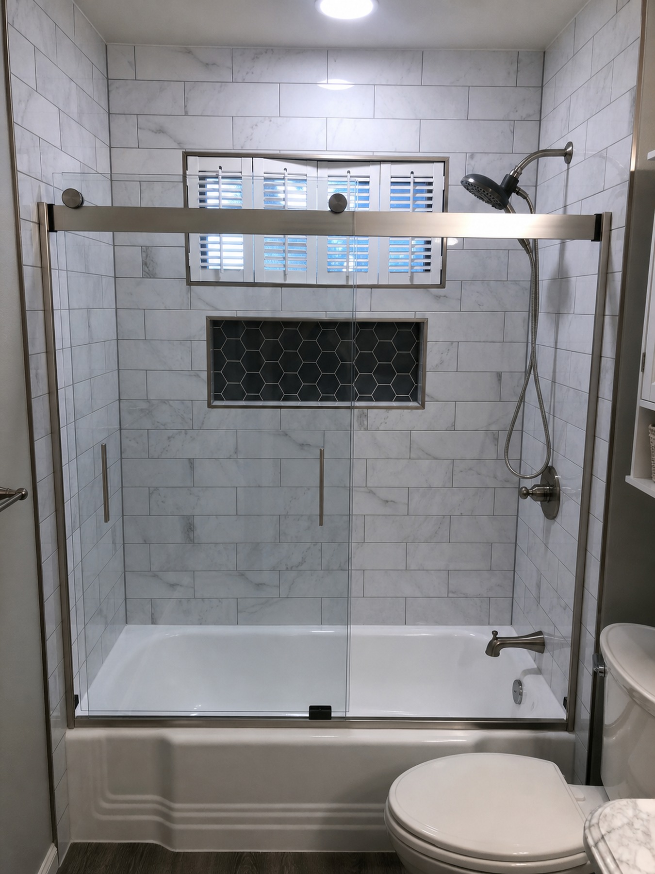 Finished tub shower with glass door, gray tile surround, and recessed niche