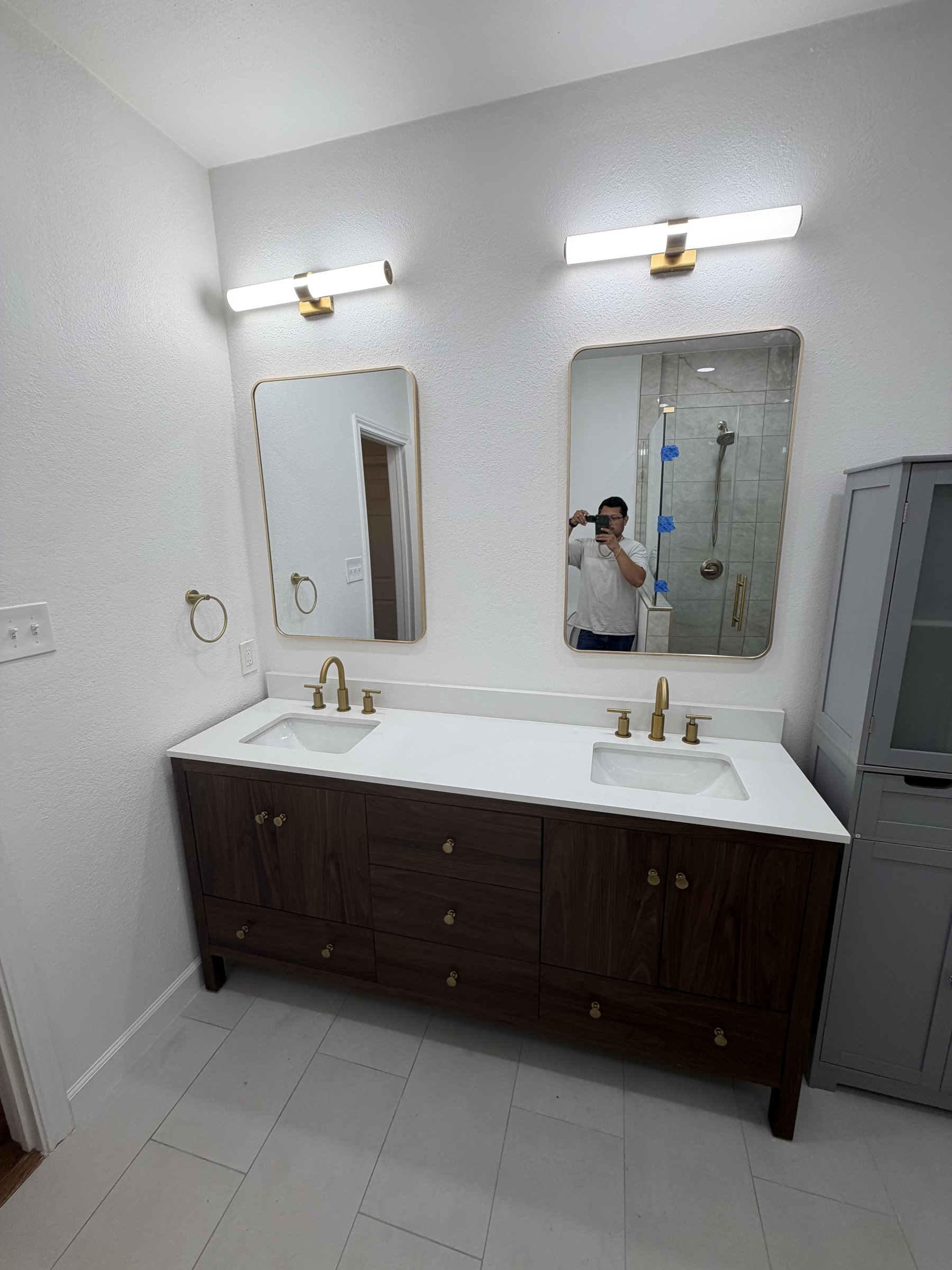 New dark wood vanity with brass fixtures, gold-framed mirrors, and gray linen cabinet